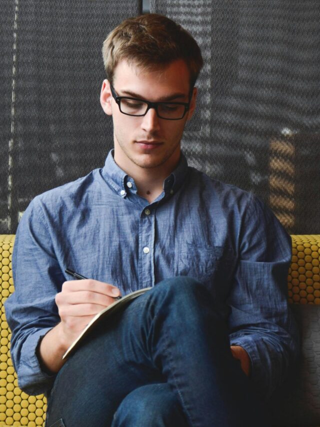A young man in glasses writes in a notebook while sitting on a stylish couch indoors.