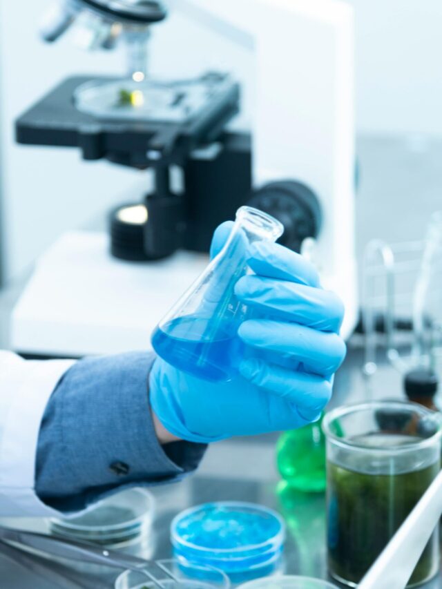 Scientist in gloves analyzing blue liquid in a laboratory setting with microscope and glassware.