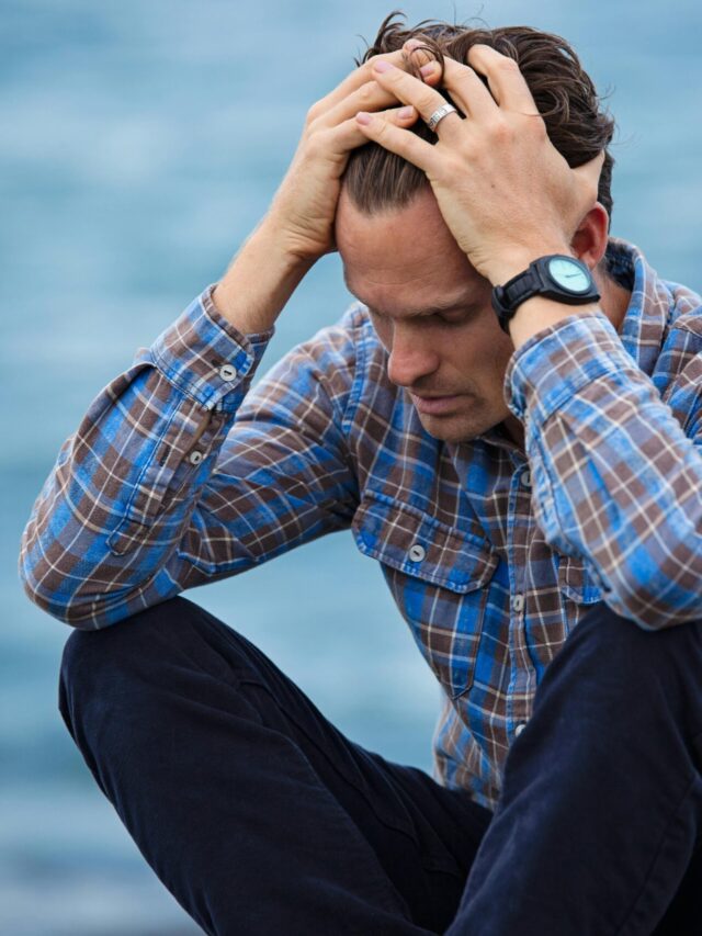 A man in a plaid shirt sits by the water looking distressed, symbolizing stress.