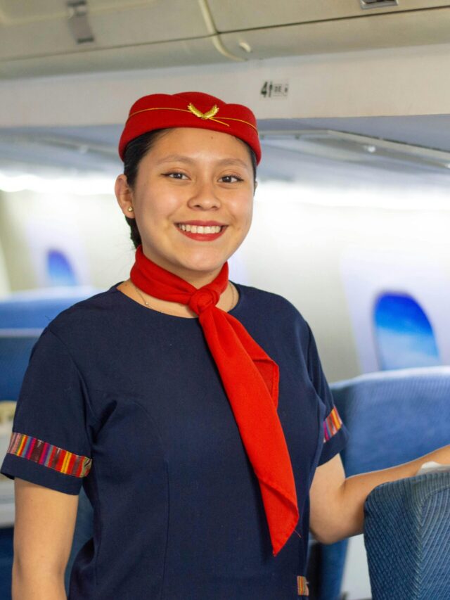 Enthusiastic flight attendant wearing a red uniform stands smiling in an airplane cabin.
