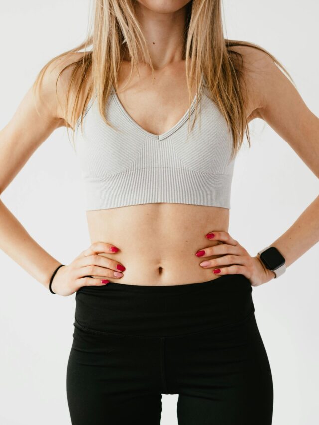 Faceless slim anonymous blond female in sports bra and black leggings in wearable bracelet showing perfect belly on white background while standing with hands on waist