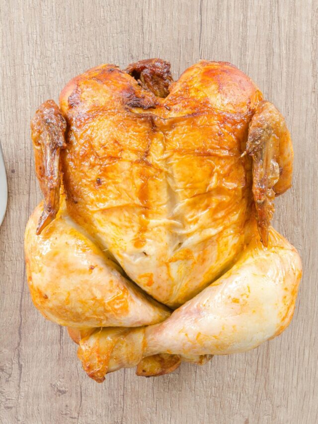 Top view of a perfectly roasted chicken placed between fork and knife on a wooden table.